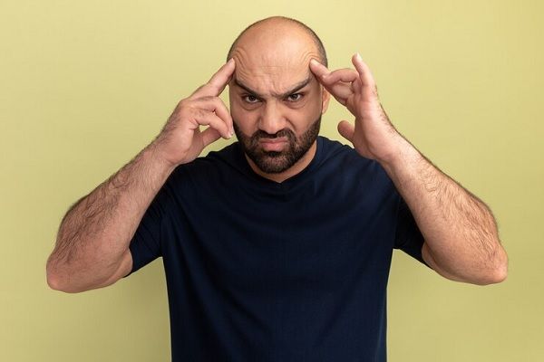 Homme barbu en t-shirt noir qui a l'air bouleversé et bouleversé de toucher sa tête souffrant de graves maux de tête debout sur un mur vert