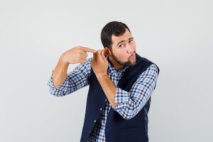 Jeune homme montrant son oreille tirée par le doigt en chemise, gilet, vue de face. perte auditive