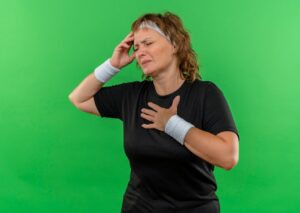 Femme sportive d'âge moyen en t-shirt noir avec bandeau, se touchant la tête, se sentant malade et fatiguée, debout sur un mur vert. 