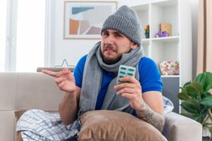 Jeune homme malade et irrité, vêtu d'une écharpe et d'un bonnet d'hiver, assis sur un canapé dans le salon, un oreiller sur les jambes, tenant un paquet de gélules, regardant droit devant lui et pointant sur le côté. la grippe