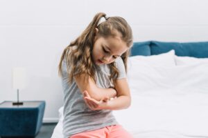 Portrait d'une jeune fille assise sur un lit blanc et regardant son coude blessé.