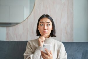 Portrait d'une jeune fille assise sur un canapé avec un smartphone, l'air pensif et hésitant devant un téléphone portable.