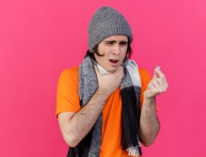 Un jeune homme malade et faible, vêtu d'un bonnet et d'une écharpe d'hiver, tient une pastille et s'agrippe à son mal de gorge, isolé en rose. 