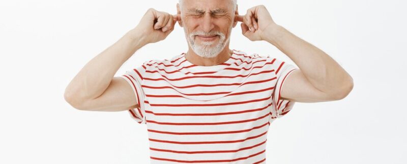 Portrait d'un grand-père mécontent qui se bouche les oreilles avec ses doigts et grimace au bruit. L’otite