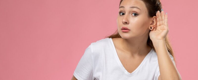 Vue de face d'une jeune femme séduisante en t-shirt blanc posant et essayant d'entendre sur un fond rose. Perte auditive