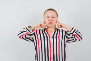 Jeune femme en blouse rayée se bouchant les oreilles avec ses index et fermant les yeux, l'air contrarié.