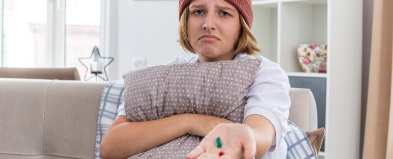 Jeune femme malade avec un bonnet chaud et une couverture, se sentant mal et souffrant du froid et de la grippe, tenant un oreiller et des pilules, inquiète, assise sur un canapé dans un salon lumineux. Qu’est-ce que la grippe
