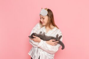 Jeune femme en pyjama et masque de sommeil posant avec un chaton gris sur fond rose.