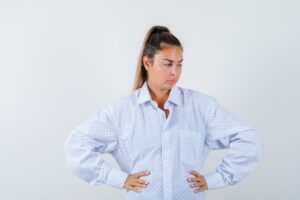Jeune femme les mains sur les hanches, réfléchissant à quelque chose, vêtue d'une chemise blanche et l'air pensif.
