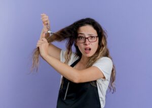 Jeune coiffeuse professionnelle en tablier tenant des ciseaux et coupant les cheveux, l'air confus.