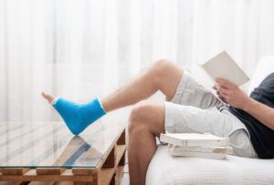 Un homme à la jambe cassée et plâtrée lit des livres sur un fond clair de l'intérieur.