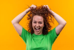 Jeune femme amusante aux cheveux courts et bouclés, vêtue d'un t-shirt vert, regardant l'appareil photo, excitée et heureuse de toucher ses cheveux.